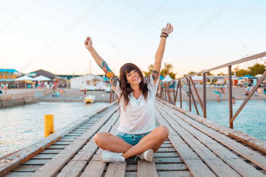 A pretty woman with tattoos sits cross-legged on a wooden pier by the sea, her hands raised in joy. The concept of summer vacation.
