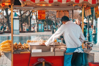 18.12.2019, İstanbul, Türkiye. Geleneksel sokak yemekleri. Mısır satıcısı elinde sigarayla tezgahın arkasında duruyor. Arka görünüm.