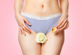 Eine Frau in ihrer Unterwäsche hält eine weiße Rosenblüte zwischen ihren Beinen auf Höhe ihrer Geschlechtsorgane und ein welkes Blütenblatt in ihrer Hand. Aus nächster Nähe. Rosa Hintergrund. Konzept der Wechseljahre und Gesundheitsprobleme von Frauen.
