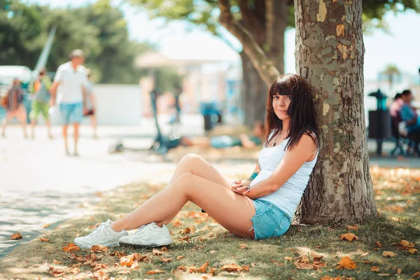 Summertime. A smiling woman is sitting relaxed leaning against a tree trunk. Recreation in the Park.