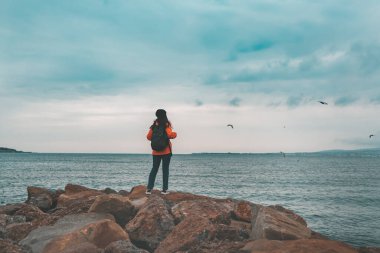 Turuncu ceketli ve şapkalı bir kadın kıyı kayalarının üzerinde duruyor. Arka planda, deniz, ufuk çizgisi ve bulutlu gökyüzü. Sonbahar. Boşluğu kopyala.
