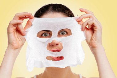 Kafkasyalı genç bir kadının portresi. Elinde bez bir maske var ve öfkeyle ona bakıyor. Beyaz arka plan. Mekanın bir kopyası. Güzellik, su ve kendine bakım kavramı..