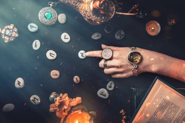 Astroloji ve ezoterizm. Cadının dişi elleri parmağıyla Runik Taşı işaret ediyor. Siyah arka planda fal, tılsım, bakır lamba ve mum bulunur. Havada ateş kıvılcımları var. Boşluğu kopyala.