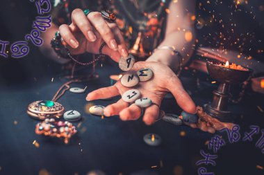 Astroloji ve ezoterizm. Cadı, mumun yanında duran rün taşlarını eline verir. Havada ateş kıvılcımları var. Karanlık arka plan. Köşelerde burç falı.