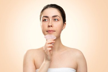 Gua Sha Masajı. Beyaz banyo havlusu giymiş bir kadın çenesine bir kuvars gua Sha kazıyıcısı ile masaj yapıyor. Bej arka plan. Uzayı kopyala Alternatif tıp ve kozmetoloji kavramı.