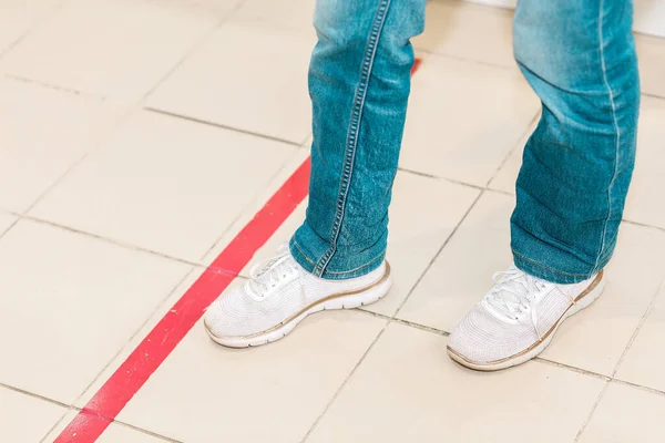 Person stand in red line, legs close-up. Attention line on the floor of the store to maintain social distance. Concept of the coronavirus pandemic and prevention measures.