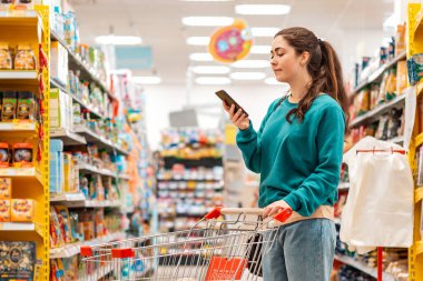 Günlük giysiler içinde genç, beyaz bir kadın market arabasını itiyor ve cep telefonu kullanıyor. Uzayı kopyala Alışveriş ve yiyecek ve iletişim konsepti.