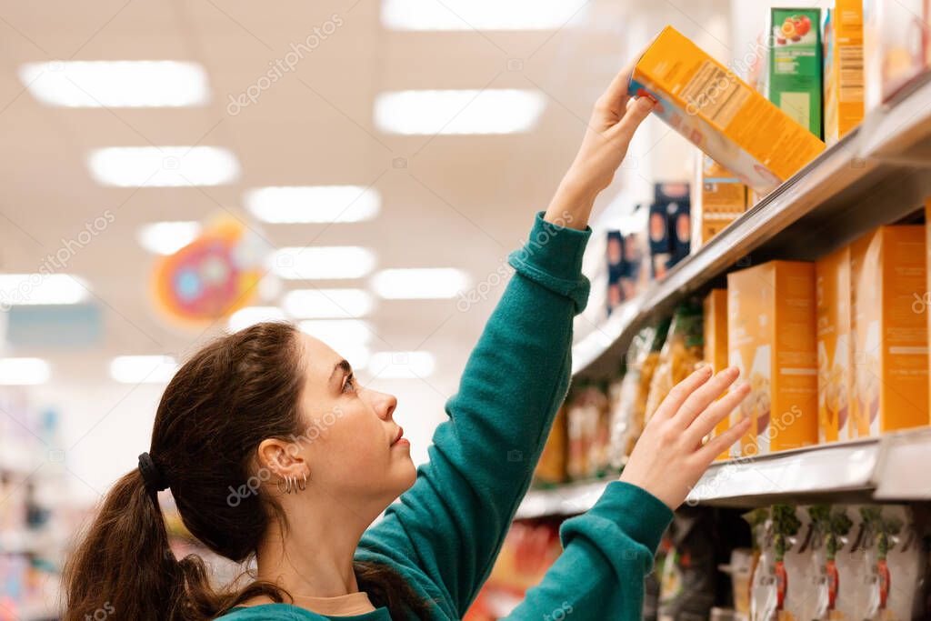 Portrait of a young Caucasian beautiful woman who reaches to the top shelf for a product. Shelves with goods in a blur in the background. The concept of buying goods and shopping.