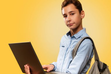 Modern teknoloji ve eğitim kavramı. Mavi gömlekli ve sırt çantalı, elinde dizüstü bilgisayar olan mutlu bir beyaz çocuk. Sarı arka plan. Boşluğu kopyala ve kapat.