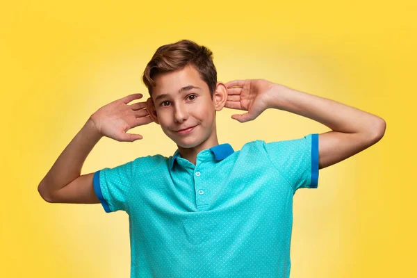 The concept of adolescence and emotions in adolescents. A teenage boy in a blue t-shirt, cheerfully pulling his ears with his hands. Yellow background. Copy space.