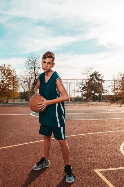 Sports and basketball. A young teenager in a blue tracksuit poses with a basketball in his hands. In the background, a basketball court. Vertical. Copy space.