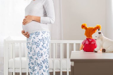 Hamile bir beyaz kadın çocukların beyaz odasında duruyor ve karnına sarılıyor. Arka planda, büyük bir pencere ve bir beşik. Kapat..