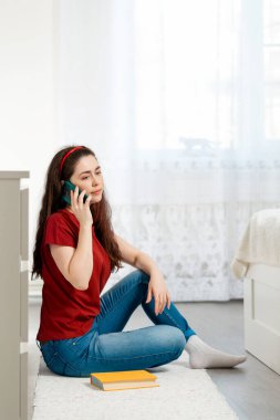 Genç bir kadın yerde oturuyor ve telefonda konuşuyor. Yanında bir de kitap var. Modern teknoloji ve iletişim kavramı.