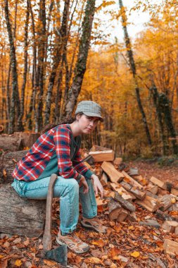Isınma mevsimi için hazırlık. Genç ve güzel bir kadın elinde baltayla kütüğün üzerinde oturuyor. Arka plandaki sonbahar ormanında. Dikey yönelim.