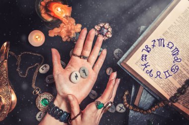 Astroloji ve ezoterizm. Cadı elinde rün taşlarını tutuyor. Siyah bir zemin üzerinde fal taşı, bir kitap, değerli tılsımlar, bakır bir lamba ve bir mum yatıyor. Kitapta Zodiac burcu var..