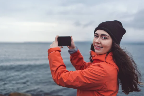 Travel and technology. A smiling woman in a hat and orange jacket takes a picture on her phone. In the background, cloudy sky and sea. Autumn. Copy space.