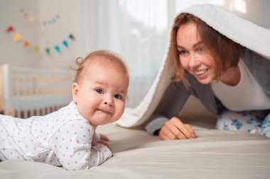 Bir battaniyenin altına saklanıp bir bebekle oynayan gülümseyen bir annenin portresi. Mutlu annelik kavramı.