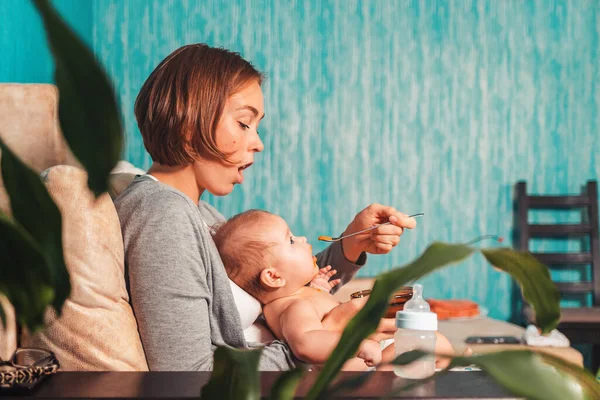 Portrait of a mother sitting on the sofa with a baby, and feeding it with a spoon. Side view. The concept of feeding and weaning baby from the breast.