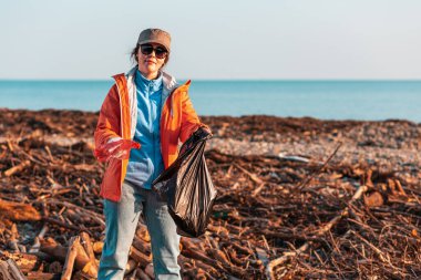 Ceketli beyaz bir kadın gönüllü çöp torbasına plastik bir şişe koyar. Arka planda, deniz ve gökyüzü ve fırtınadan sonra çamurlu bir plaj. Çevre kirliliği kavramı.