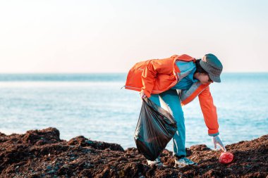 Ceketli genç beyaz bir kadın plajda plastik bir torba içinde çöp topluyor. Arka planda, deniz ve gökyüzünün ufku. Boşluğu kopyala ve kapat.