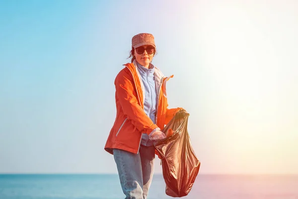A young Caucasian woman in a jacket collects garbage and puts it in a plastic bag. In the background, the horizon of the sea and sky. Copy space and light.