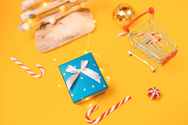 Holiday shopping. A toy shopping cart, gift box, and a craft wooden Christmas tree. Yellow background with candys and christmas balls. Top view. Concept of Christmas sales.