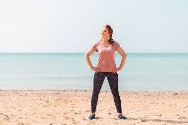 Yaz. Spor kıyafetleri içinde, elleri belinde sahilde poz veren güzel bir kadın. Arka planda deniz ve mavi gökyüzü. Boşluğu kopyala.