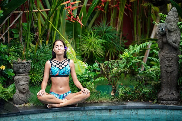 Yoga. A young woman in a bathing suit, meditating by the pool, closing her eyes. In the background, tropical plants and statues of gods. The concept of a healthy lifestyle and energy.