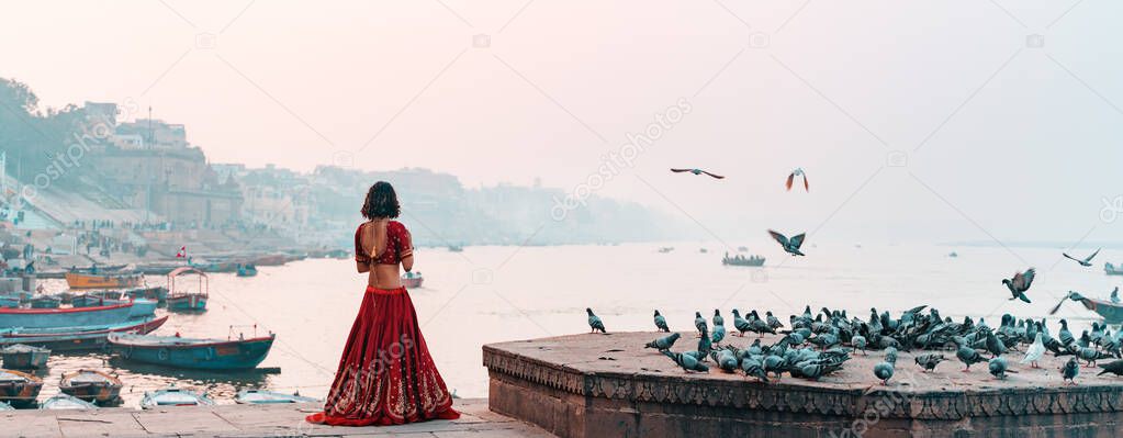 A beautiful Indian woman in a red Sari stands alone on the street. There is a flock of pigeons on the pedestal. In the background is a river and a view of the city. Travel and culture. Panorama.