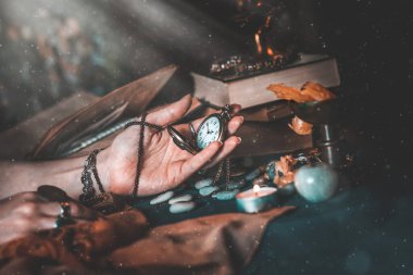Astroloji ve ezoterizm. Cadının dişi eli zincirli bir saat tutuyor. Arka planda eski kitaplar, fal işaretleri ve bir mum var. Işık ve toz.