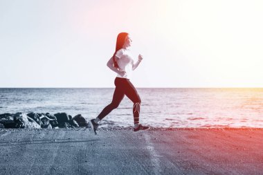 Spor ve deniz boyunca koşu. Genç esmer bir kadın kayalık bir sahil boyunca bir yol boyunca koşar. Arka planda, deniz ve gökyüzü. Tek Renkli.