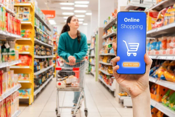 一个人的手拿着一部手机 屏幕上有一个购物篮 在背景中 一个女人在超市推着一辆购物车 看起来很模糊 网上购物的概念 — 图库照片
