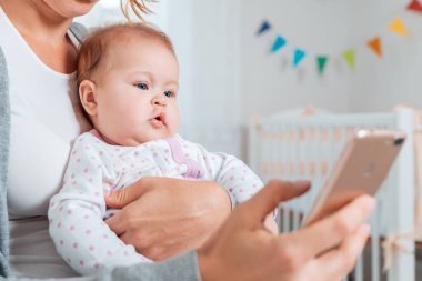 Annesinin kollarında oturan ve kullandığı akıllı telefona bakan bir bebeğin portresi. İnternet 'i kullanarak çocukları eğlendirme ve eğitme kavramı.