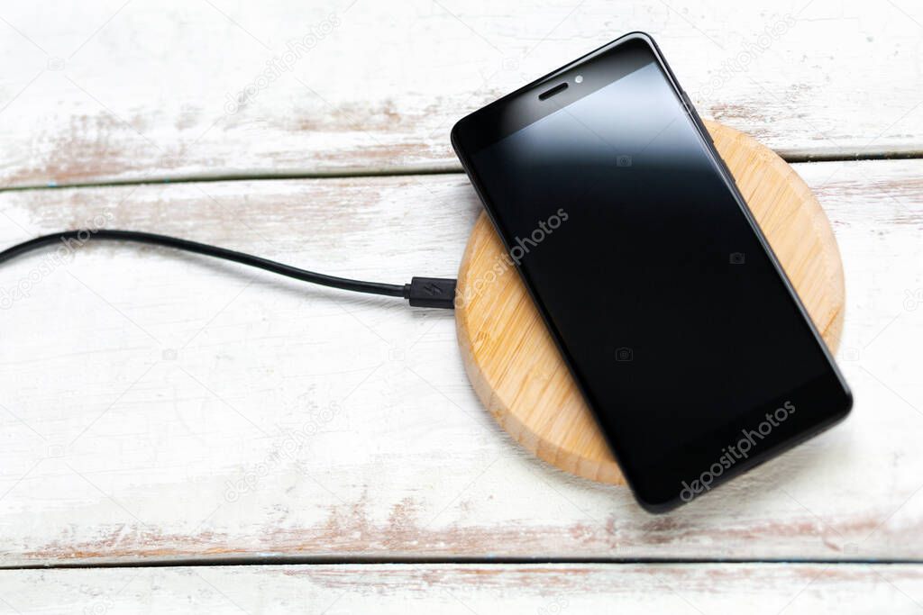 Concept of modern technologies. The black smartphone is on a wireless charger. White wooden background with horizontal stripes. Copy space. Flat line.