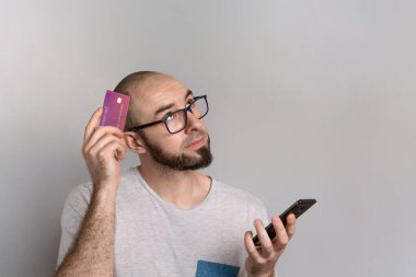 Bankacılık ve kredi kartı harcamaları kavramı. Gözlüklü bir adam bir elinde banka kartı, diğer elinde cep telefonu tutuyor. Düşüncelilik duygusuyla. Boşluğu kopyala.