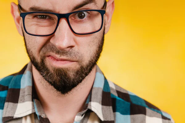 People and emotions. A bearded man wearing glasses and a blue checked shirt expresses his dislike. Yellow background. Close up.