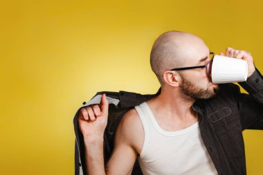 Sabah uyanışı ve verimlilik kavramı. Kel, sakallı, gözlük takan, gömlek giyen ve fincandan kahve ya da çay içen bir adam. Sarı arka plan. Boşluğu kopyala.