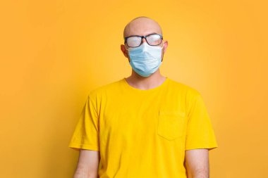 Virüs koruması. Sarı arka planda sarı tişörtlü, sis gözlüklü ve tıbbi maskeli kel bir adam duruyor. Karantina, kendini izole etme ve koronavirüs kavramı.
