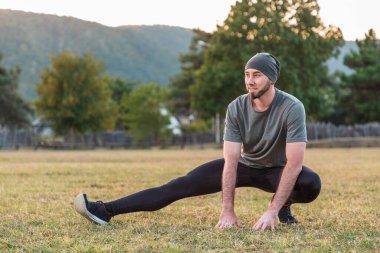 Spor ve yoga. Spor giyim kuşamında sakallı bir adam antrenmandan önce çimlerin üzerinde gülümsüyor ve esneme hareketleri yapıyor..