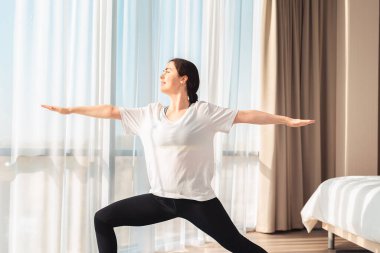 Yoga. Genç ve güzel bir kadın evde antrenman yapıyor, savaşçı pozu veriyor. Yan görüş. Arka plandaki pencereler. Ev egzersizi konsepti.