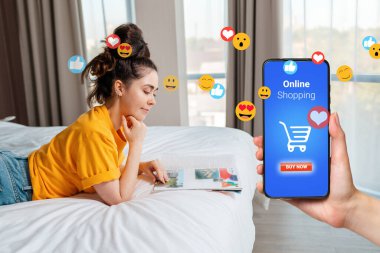 İnternetten alışveriş. Genç, gülümseyen bir kadın yatakta uzanıyor ve bir dergi okuyor. Akıllı telefonlu, market arabalı ve uçan emojili. Yan görünüm.