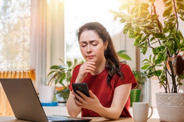 Serbest. Genç bir kadın akıllı bir şekilde kullanır. Ev dekorasyonu, pencerenin dışındaki güneş, bir dizüstü bilgisayar ve masada bir fincan çay. Karantina, kendini izole etme ve uzaktan çalışma kavramı..