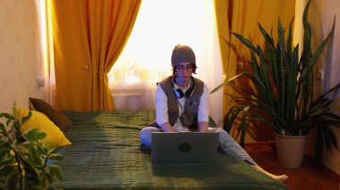 Şık giyinmiş, gözlüklü beyaz bir kadın yatağında dizüstü bilgisayarla yazı yazıyor, evden ve modern online iş hayatı tarzından uzak çalışma kavramı var. Geniş açı