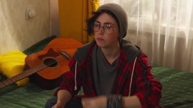 Gözlüklü genç esmerin portresi televizyon izlerken yatakta patlamış mısır yiyor ve akıllı telefon kullanıyor, gençlik eğlencesi ve dijital alışkanlık kavramı.