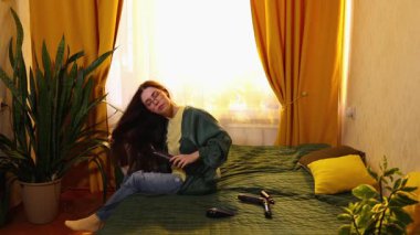 Koyu kıvırcık saçlı beyaz kadın, saç kurutma makinesini rahat bir odada saç şekillendirme, günlük kişisel bakım ve sağlık konsepti için kullanıyor. Geniş açı