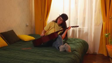 Genç beyaz kadın evde yatağında gitar çalarak ve müzik öğrenerek sanatsal becerilerini ve konsantrasyonunu geliştirmeye çalışıyor..