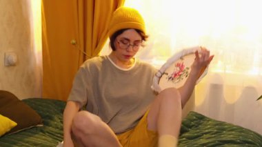 Genç bir kadın yatak odasında, çemberle nakış işliyor çapraz dikiş teknikleri çalışıyor ve sakin ve rahat bir atmosferde vakit geçiriyor. Orta çekim.