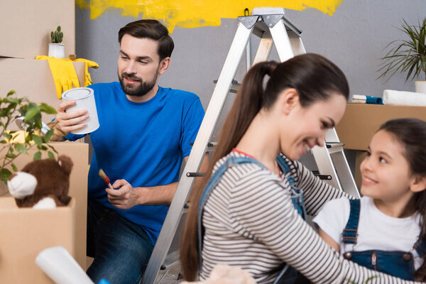 мать обнимает дочь, в то время как отец держит кисть и краски банка, семья делает ремонт в новой квартире
 