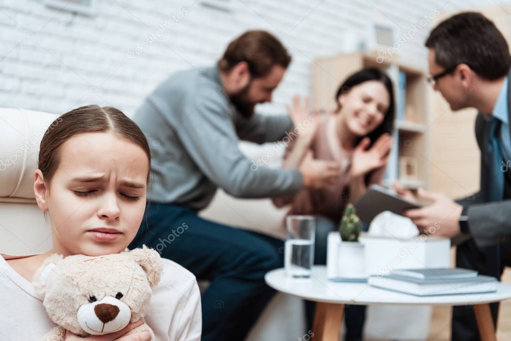 Little girl with teddy bear in hands avoiding scandal of parents in psychiatrists office