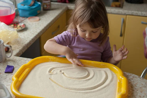 Little girl draws a finger on a tray with sand in the kitchen, make fine motor skills with drawing